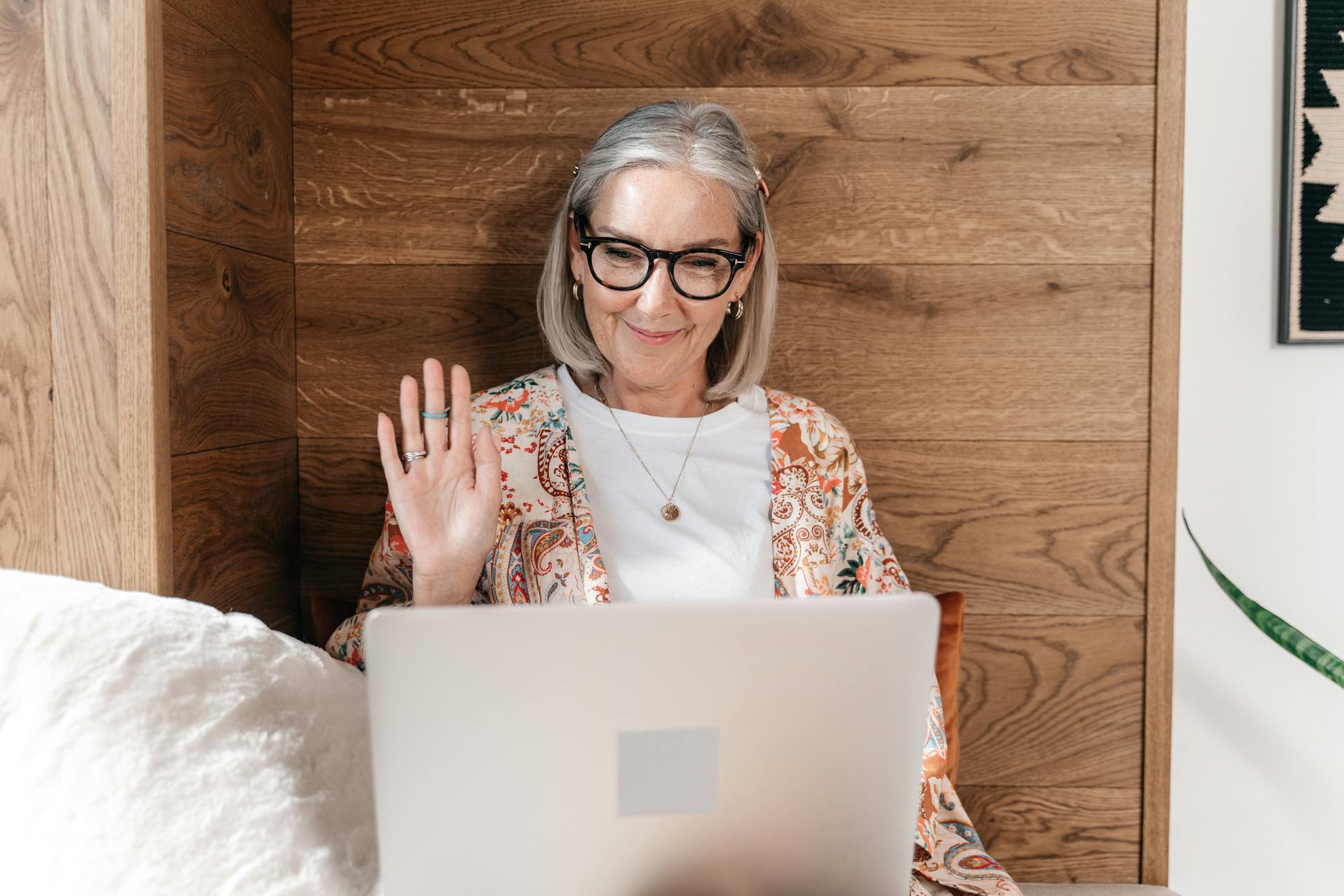 Senior woman with gray hair and glasses smiling and waving at her laptop during a Social Circle video call